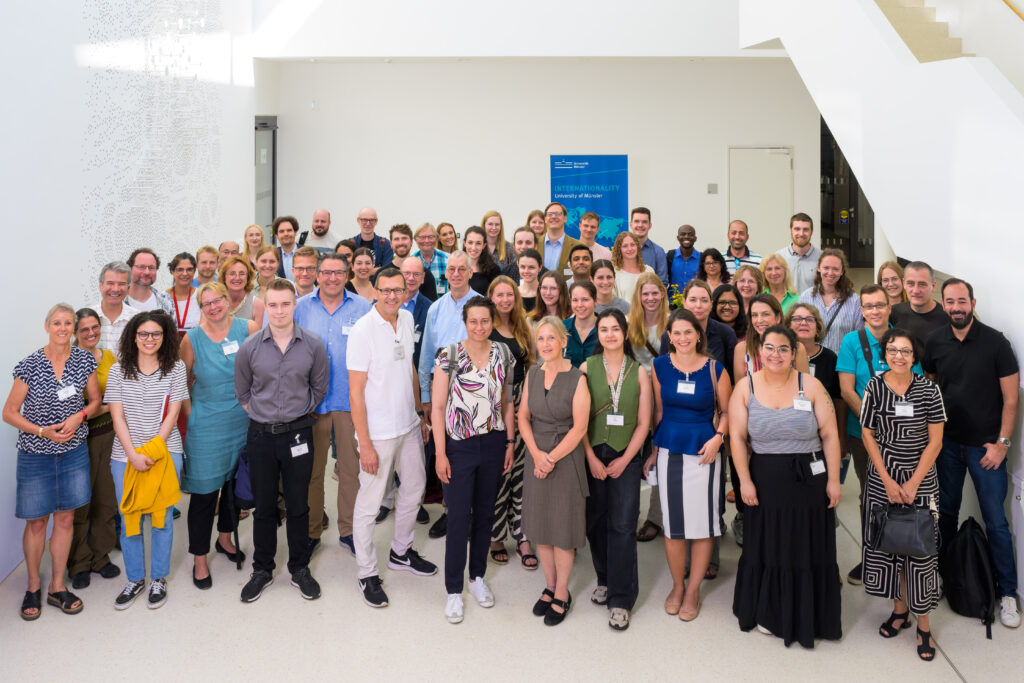 Gruppenfoto der lächelnden Symposiumsteilnehmenden im weißen Foyer des Multiscale Imaging Centres in Münster. Foto Credits: Erk Wibberg