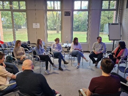 Die Teilnehmenden des ACHIEVE Mentoring-Meetings sitzen im Stuhlkreis in einem hellen Raum mit hohen Fenstern und diskutieren.