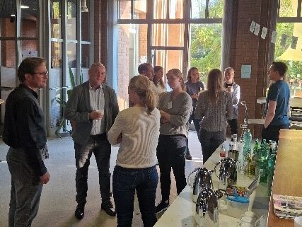 Die Teilnehmenden des ACHIEVE Mentoring-Meetings stehen in Gruppen um ein Kaffee-Buffet herum und reden miteinander.