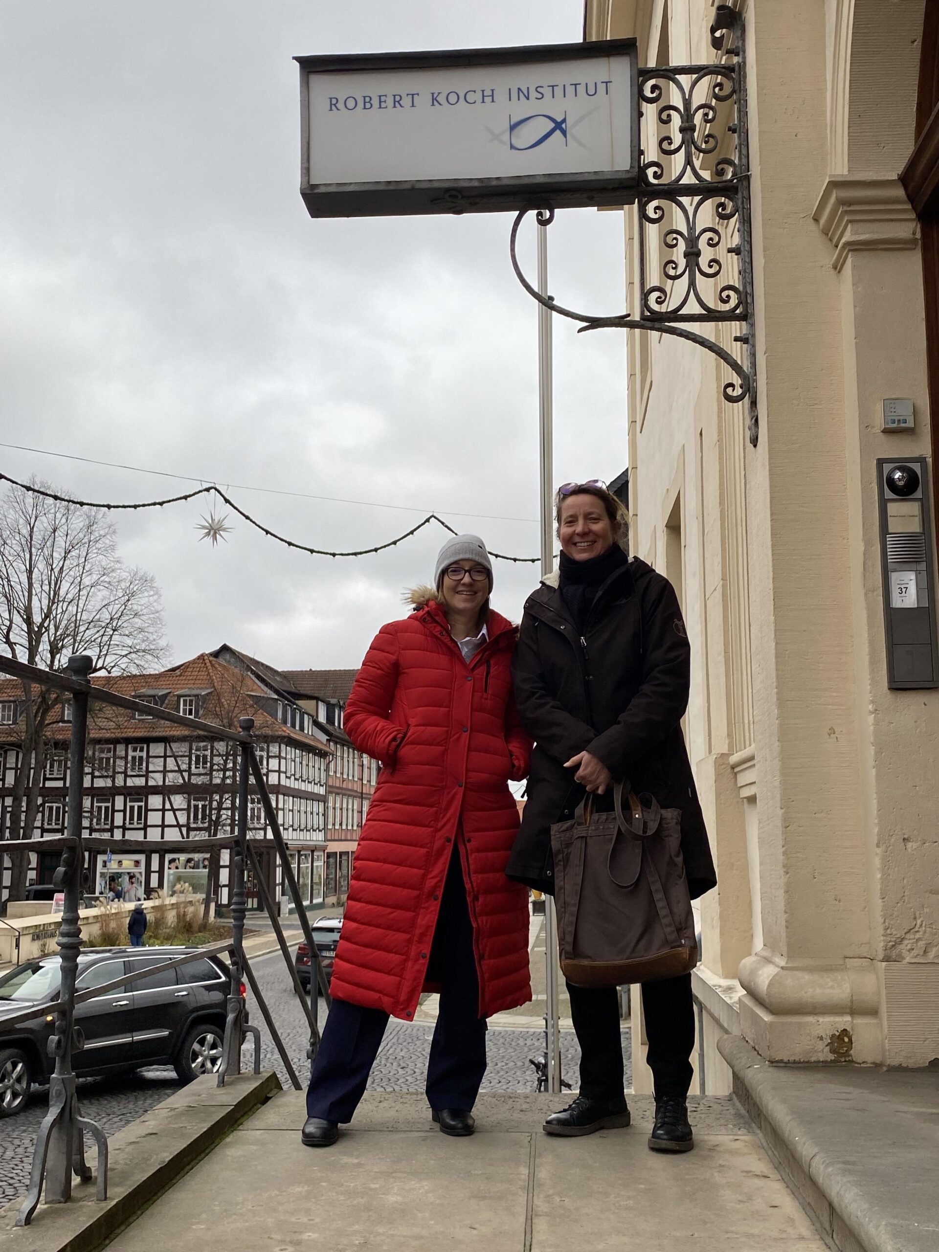 Foto: Zwei Frauen in Wintermänteln stehen auf dem Treppenaufgang zum Robert-Koch-Institut in Wernigerode. Hinter ihnen sieht man die Fachwerkhäuser der historischen Altstadt von Wernigerode. Links im roten Mantel steht Professorin Dr. Caroline Barisch, rechts daneben in dunklem Mantel und mit großer brauner Tasche steht Professorin Dr. Antje Flieger.