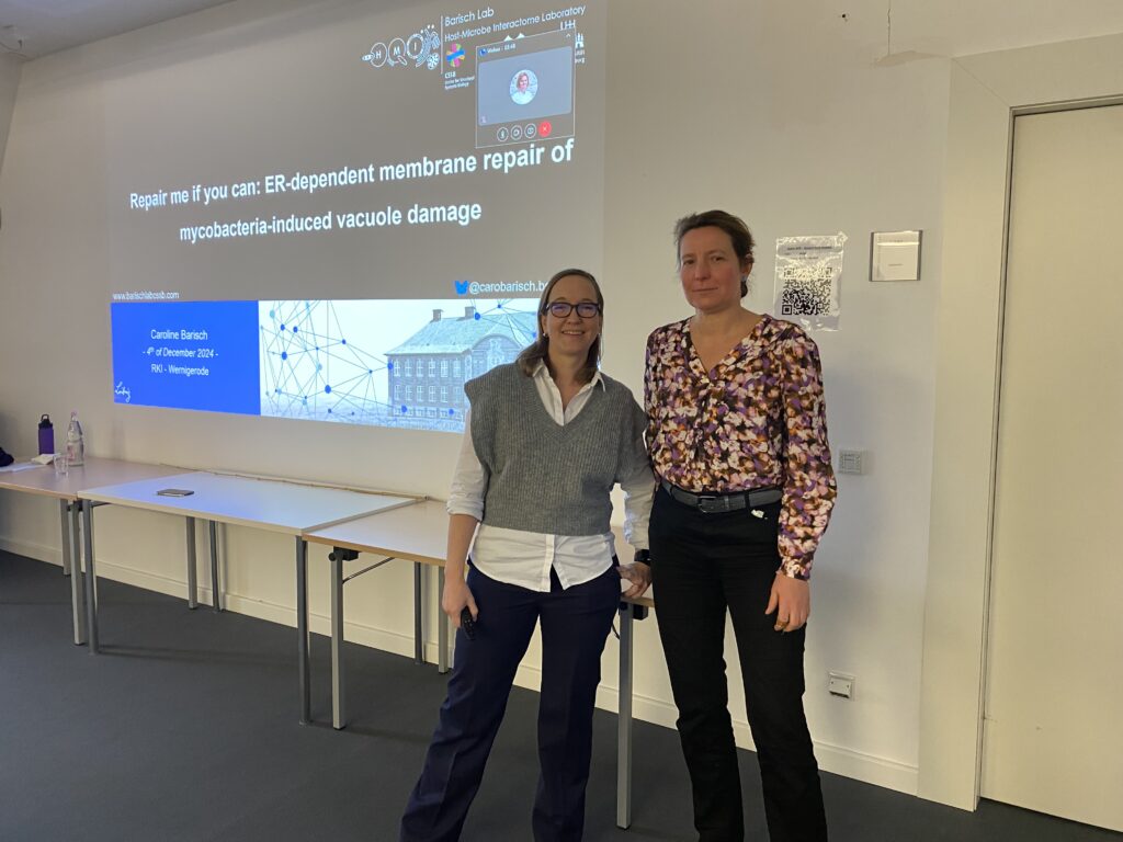 Die Mikrobiologinnen Professorin Dr. Caroline Barisch und Professorin Dr. Antje Flieger stehen vor einer Bildschirmpräsentation mit dem englischen Titel Repair me if you can: ER-dependent membrane repair of mycobacteria-induced vacuole damage.