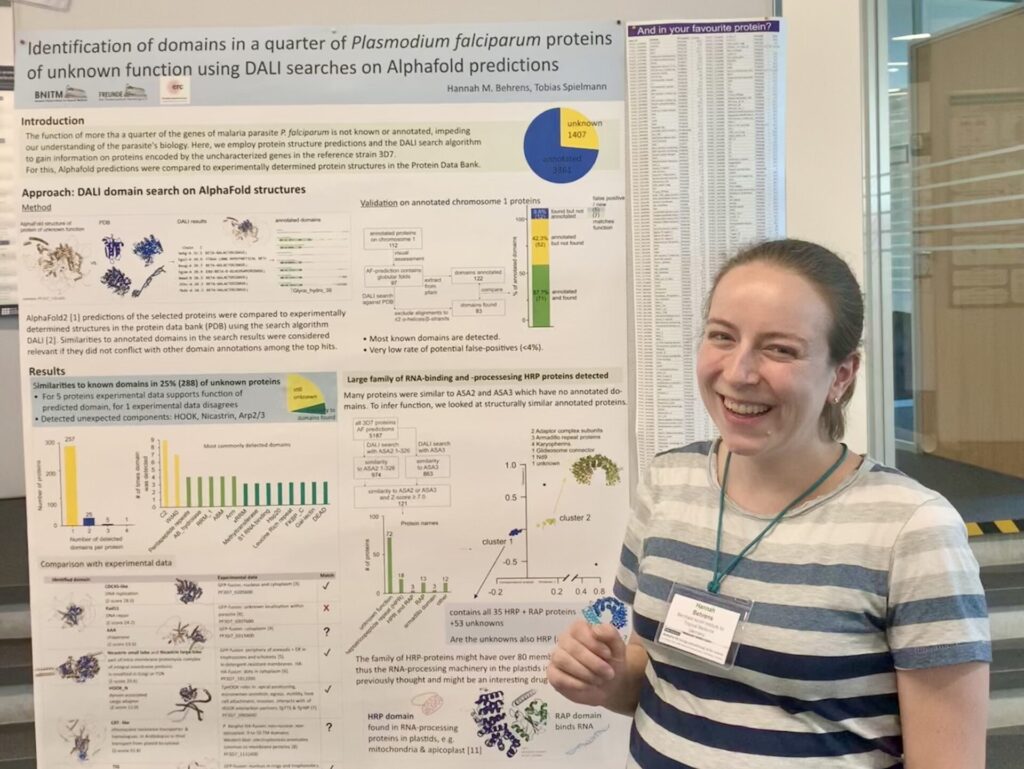 Foto: Eine junge Frau steht rechts neben einem wissenschaftlichen Poster und lacht stolz in die Kamera. Der englische Titel des Posters lautet: Identification of domains in a quarter of Plasmodium falciparum proteins of unknown function using DALI searches on Alphafold predicitions.